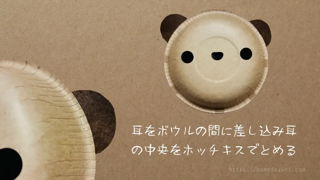 耳のパーツを紙ボウルの間に挟み、中央をホッチキスで留めている様子。「耳をボウルの間に差し込み耳の中央をホッチキスでとめる」という文字入り。