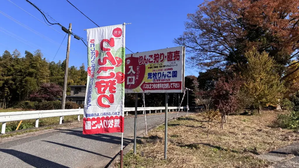「荒巻りんご園の入口看板と“りんご狩り・直売”ののぼり旗。晴天のもと、道路脇に案内が設置されている様子