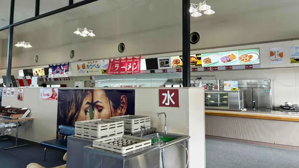 カフェテリア「カウベル」の注文カウンター。ラーメンやカレーなどのメニュー札が並び、セルフの水飲み場がある。
