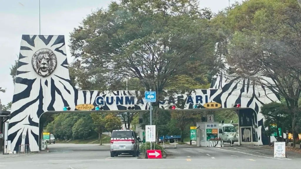 群馬サファリパークのシンボル、ゼブラ柄の大きなアーチ型メインゲート。「GUNMA SAFARI」の文字とライオンのモチーフが刻まれ、バスと乗用車の入口が左右に分かれている。