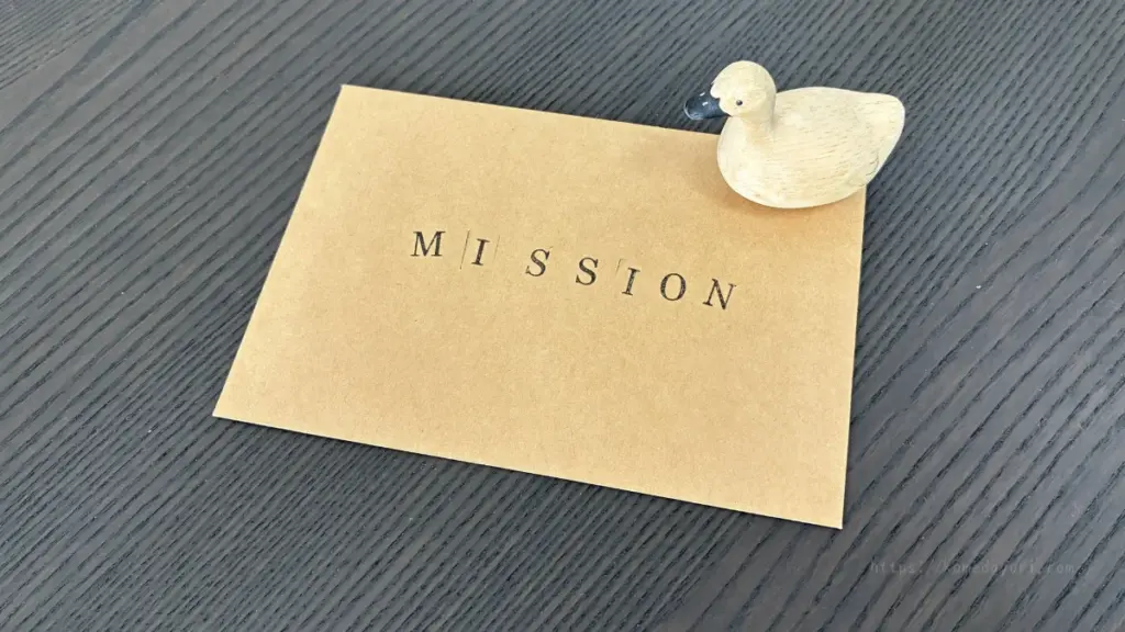 「MISSION」とスタンプされたクラフト封筒の上に木彫りのアヒルが乗っている様子。