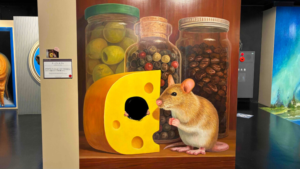 巨大チーズとネズミのトリックアートで遊ぶ子ども。チーズの穴から顔をのぞかせている。