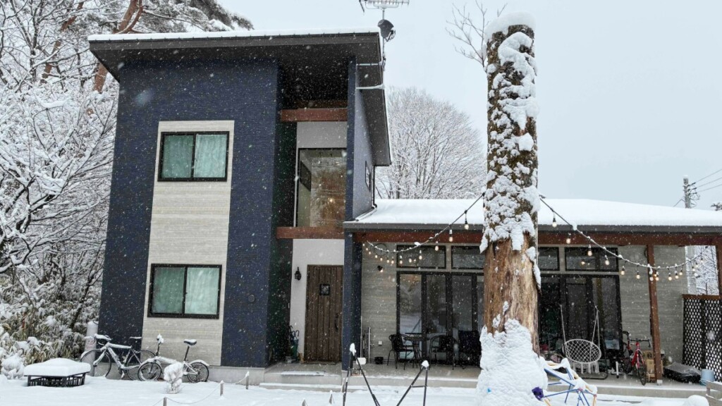 雪が降りしきる中、モダンな黒い2階建てのペンション「トロールの森ステラ」を正面から捉えた外観。手前の大きな木の幹やテラスの屋根、庭の自転車に雪が積もっており、テラスにはイルミネーションライトが灯っている。