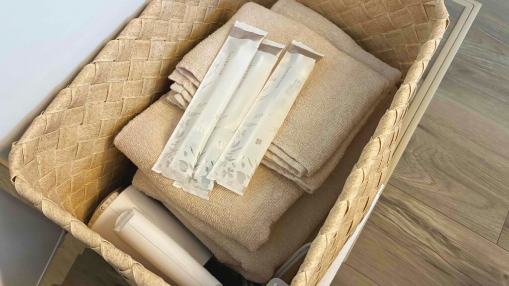 カゴの中に用意されたベージュのバスタオル、フェイスタオル、および個包装された歯ブラシセット3点。