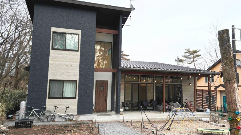 黒っぽい2階建ての建物と平屋部分が連結したモダンな一軒家「トロールの森ステラ」の外観。手前の庭には子供用のドーム型遊具やトランポリンがおいてある。