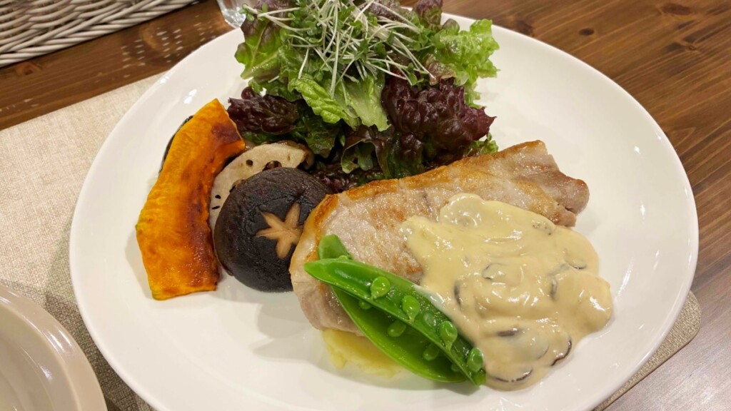 白い丸皿に盛り付けられた、キノコ入りのホワイトソースがかかった豚肉のソテー。付け合わせにカボチャ、レンコン、椎茸の素焼き、スナップエンドウ、グリーンサラダが添えられている。