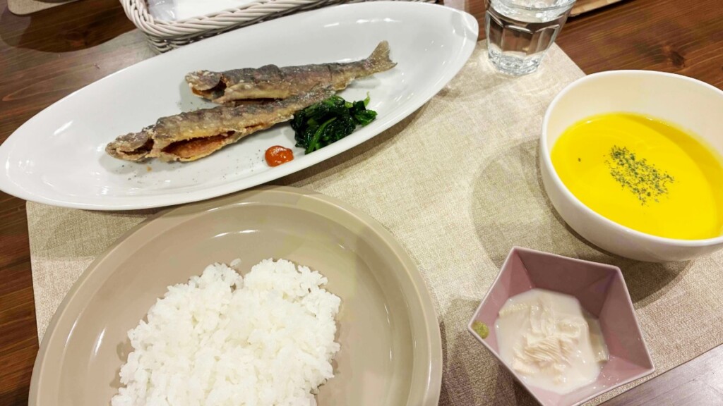 白い長皿に盛り付けられた2尾の魚の唐揚げとほうれん草、お椀に入った黄色いポタージュスープ、小鉢に盛られた白い湯葉、茶碗に盛られた白米が置かれている。