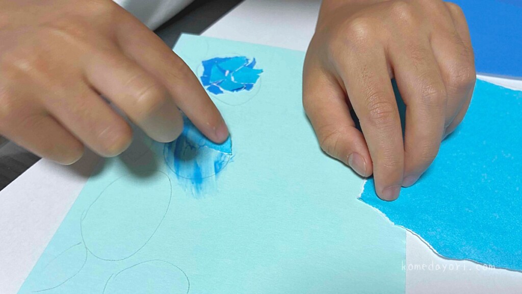 子どもが青い折り紙をちぎって紫陽花の下書きにのりで貼っている様子